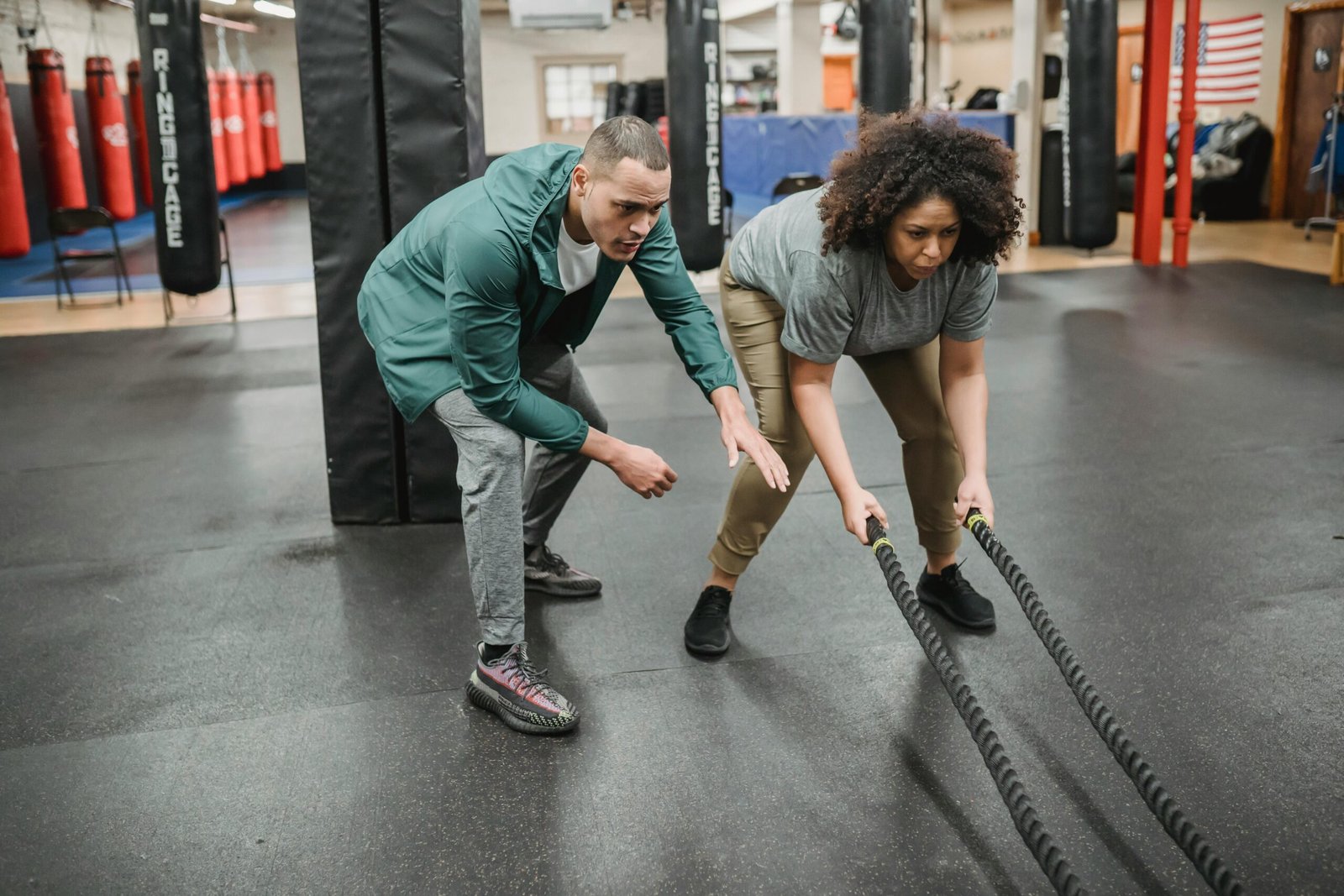 Home Full body focused African American female in activewear exercising with battle ropes near professional male instructor in contemporary fitness center